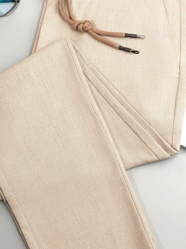 The Pleated Trouser in stone cotton–linen basketweave, laid flat on grey pattern paper.